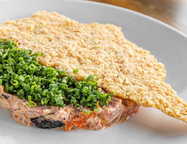 A savory open-faced toast topped with chopped chives, on a slice of meat with a crispy rice cracker perched beside it.