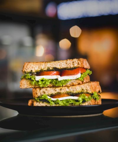 A stacked sandwich/burger with lettuce, tomato, cucumber, and cheese on a plate, with bokeh lights blurred in the background.