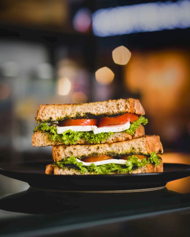A stacked sandwich/burger with lettuce, tomato, cucumber, and cheese on a plate, with bokeh lights blurred in the background.