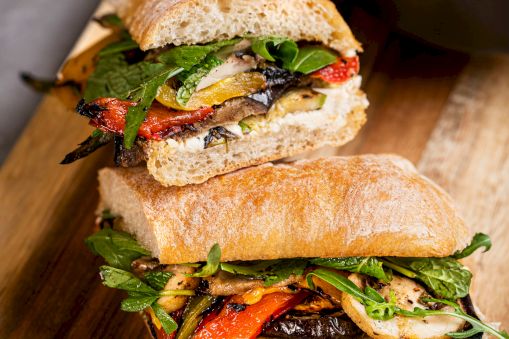 A grilled veggie sandwich with crusty bread, leafy greens, roasted peppers, mushrooms, and melted cheese, ready to eat.