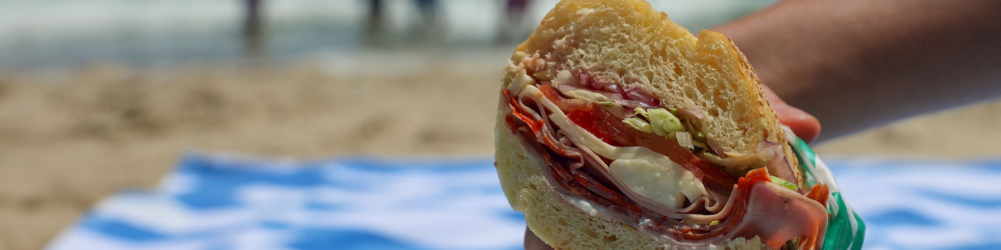 A hand holds a deli-style sandwich with veggies on a seaside beach, with people and ocean blurred in the background.