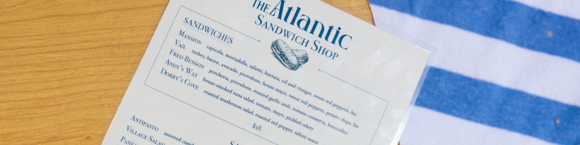 A &ldquo;The Atlantic&rdquo; sandwich menu and green-and-white wrapped sandwiches sit on a wooden board beside blue-and-white striped cloth.