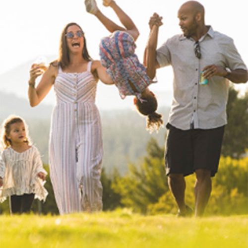 A family of four is having fun outdoors. The parents are holding a child upside down while another child walks beside them in a grassy area.