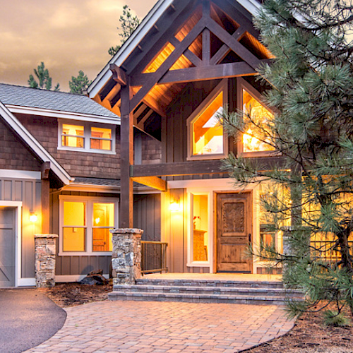 A large, modern house with multiple stories, warm exterior lighting, a driveway with stone pavers, and surrounded by trees at dusk.