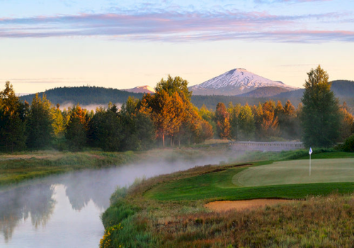 A serene landscape featuring a golf course beside a reflective body of water, surrounded by trees, with a snow-capped mountain in the background.