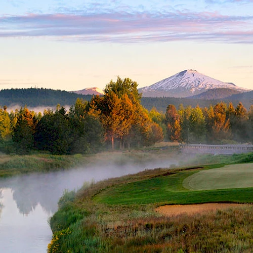 A serene landscape featuring a golf course beside a reflective body of water, surrounded by trees, with a snow-capped mountain in the background.