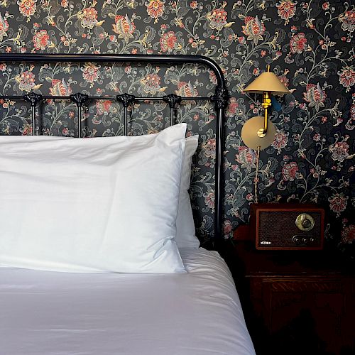 The image shows a neatly made bed with white linens, set against a floral wallpaper. There is a small nightstand with a lamp and vintage radio on it.