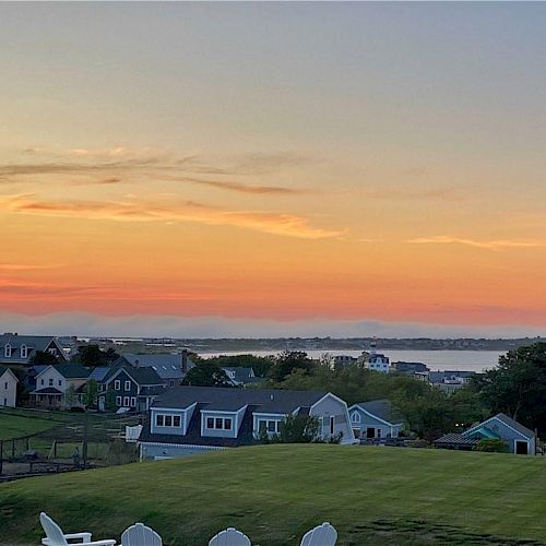 This image shows a serene sunset over a residential area with houses, a grassy lawn, and a body of water in the background.