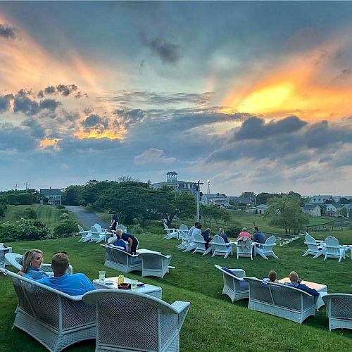 People are relaxing on a grassy area with white chairs and sofas, enjoying a vibrant sunset under a partly cloudy sky, showcasing a peaceful evening.