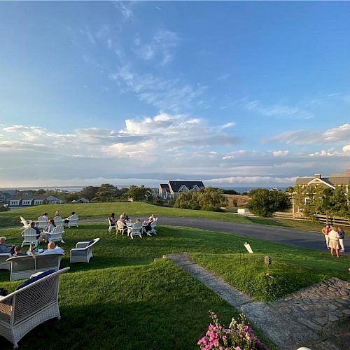 People are relaxing on outdoor furniture in a grassy area with beautiful scenic views. The sky is partly cloudy, and some houses are in the background.