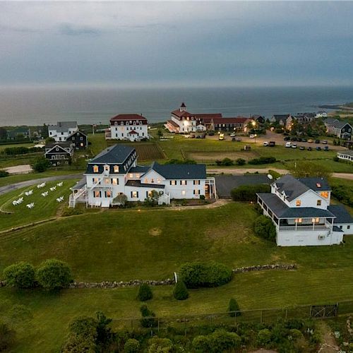 Aerial view of a scenic coastal area featuring several houses, vast green lawns, and a nearby body of water under a cloudy sky.