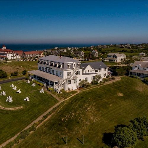 An aerial view showcases a large house on a hill, surrounded by grassy lawns with white chairs, neighboring buildings, and distant ocean views.