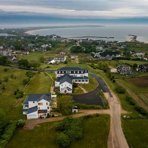 An aerial view shows houses in a coastal village with fields, roads, and greenery, ending with the sea at the horizon.