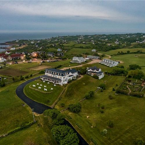 The image shows an aerial view of a coastal area with large houses, expansive lawns, and a winding road, surrounded by green fields and distant water.