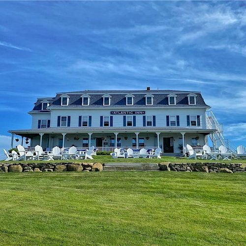 A large, multi-story building, likely a hotel or inn, with outdoor seating and a wide lawn under a clear blue sky with clouds.