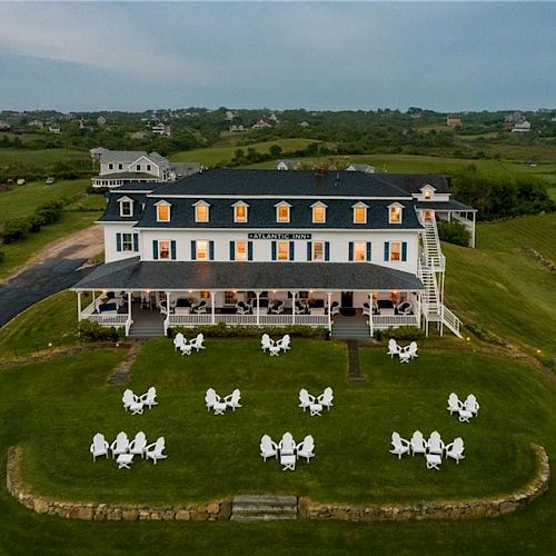 An image showing a large building with a wraparound porch and white chairs arranged on the lawn in front of it, set in a rural landscape.