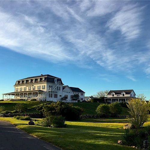 A large white house with multiple gables and a wraparound porch sits on a grassy hill, with a smaller house nearby and a partly cloudy sky overhead.