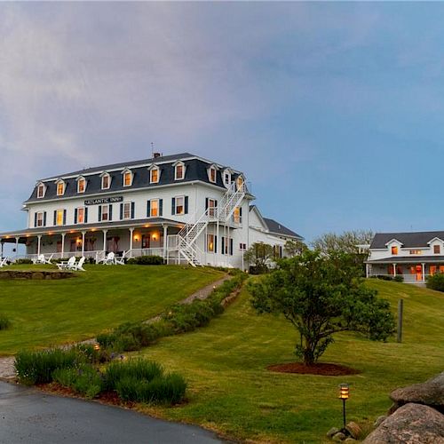 This image shows a large, elegant building resembling a historic inn or hotel on a grassy hill with surrounding landscaped gardens and a smaller building.