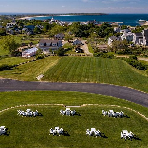 The image shows a coastal village with houses, green fields, a road, and sitting areas with chairs on a lawn, overlooking the ocean.