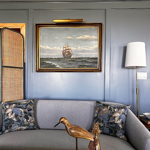 A cozy living room features a blue couch with floral cushions, a wooden bird statue, a framed ship painting, patterned wallpaper, and a floor lamp.