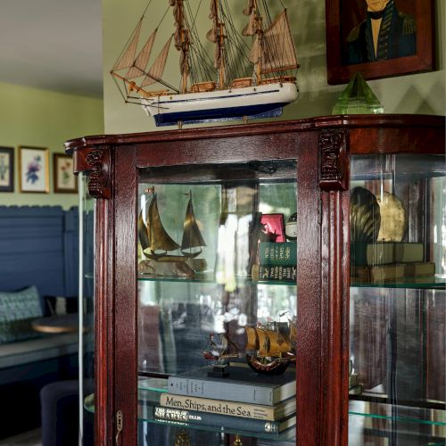 A wooden cabinet displays model ships and books; a ship model sits atop, with a portrait on the wall behind.