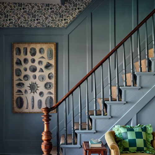 A staircase with wooden railing, blue paneling, framed art, patterned wallpaper, and a yellow chair with a green pillow.