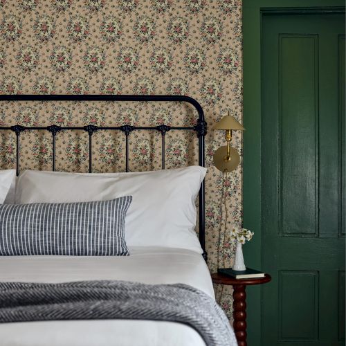 A bedroom with patterned wallpaper, iron bed, white linens, a striped pillow, a blanket, a small side table, and a green door is shown.