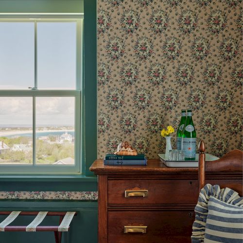 A room with a window, floral wallpaper, wooden dresser with items, a chair, and a view outside.