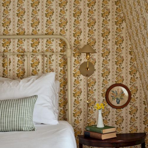 A cozy bedroom corner with floral wallpaper, metal bed frame, pillow, wall lamp, books, mirror, and a small flower vase on the nightstand.