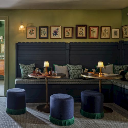 Cozy room with dark seating, small tables, round stools, and framed artwork on olive walls; ambient lighting enhances the intimate setting.