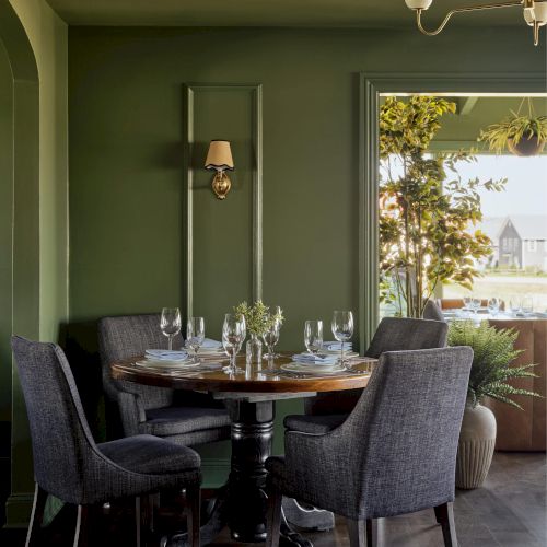 A cozy dining area with a round table, gray chairs, and elegant decor against green walls, featuring a window view of a garden scene.