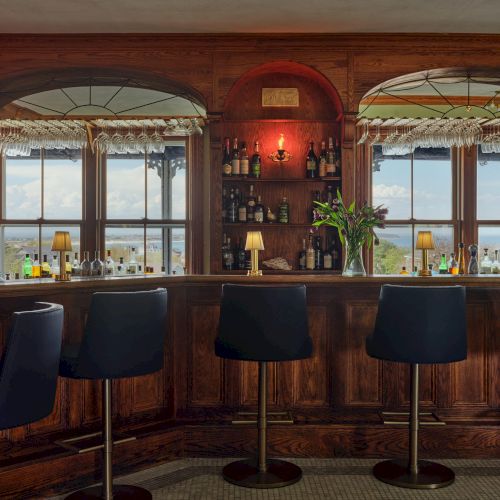 A cozy bar interior with wooden decor, stools, and shelves of bottles. Large windows offer a view outside.