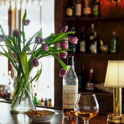 A cozy bar scene featuring purple tulips, a bottle and glass of whiskey, a bowl of snacks, and a lamp on a wooden counter.