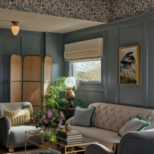 A cozy, elegant living room with a tufted sofa, armchair, plants, coffee table, panelled walls, and floral wallpaper accent.
