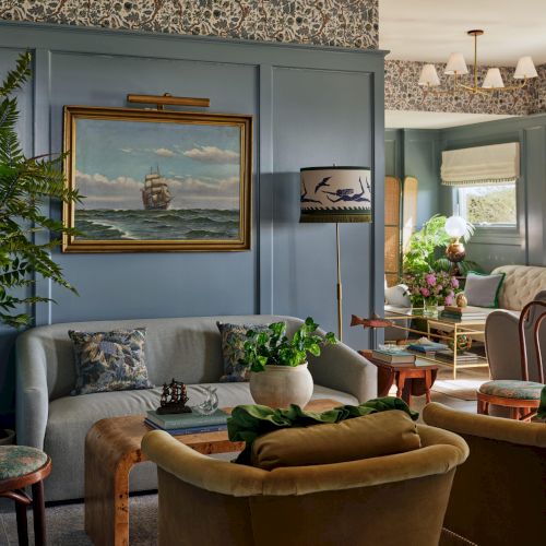 A cozy living room with blue walls, armchairs, plants, and a ship painting. A lamp and decorative wallpaper add a classic touch.