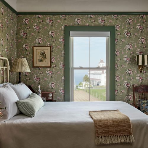 A vintage-style bedroom with floral wallpaper, a bed, and a window showing a scenic view of a lighthouse and ocean.