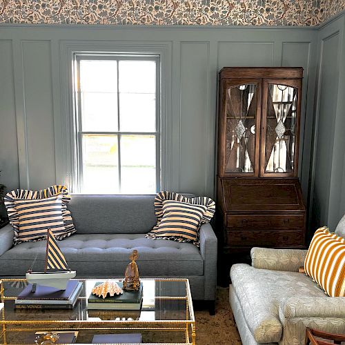 A cozy living room with blue walls, a gray sofa, patterned pillows, a glass coffee table, and a wooden cabinet. Ends the sentence.