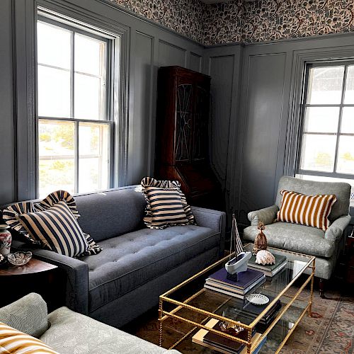 A cozy living room with gray walls, a blue sofa, striped pillows, armchairs, a glass coffee table, a wooden cabinet, and patterned wallpaper.