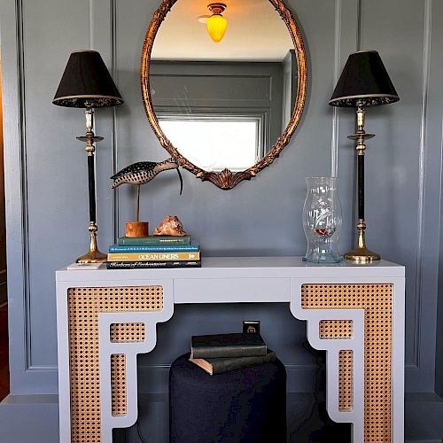 The image shows a stylish console table with two black-shaded lamps, books, a decorative sculpture, and a mirror mounted on a dark paneled wall.