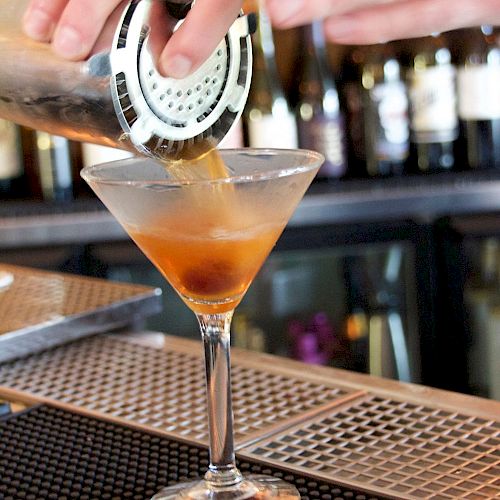 A bartender is pouring a cocktail into a martini glass using a shaker with a strainer, with a bar counter and various bottles visible in the background.