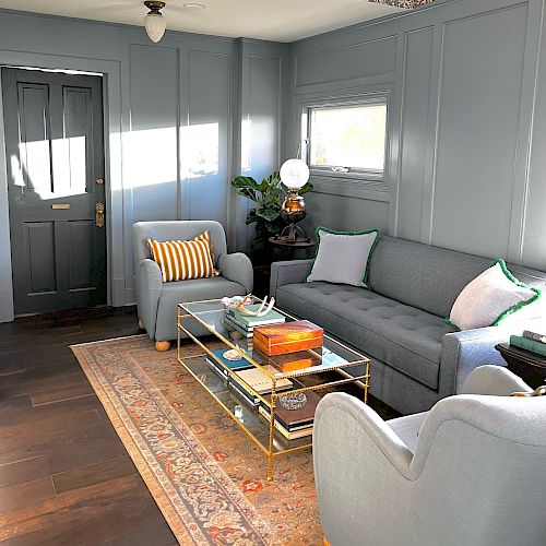 A cozy living room with gray walls features a gray couch, two armchairs, a glass coffee table with books, a rug, and a plant.