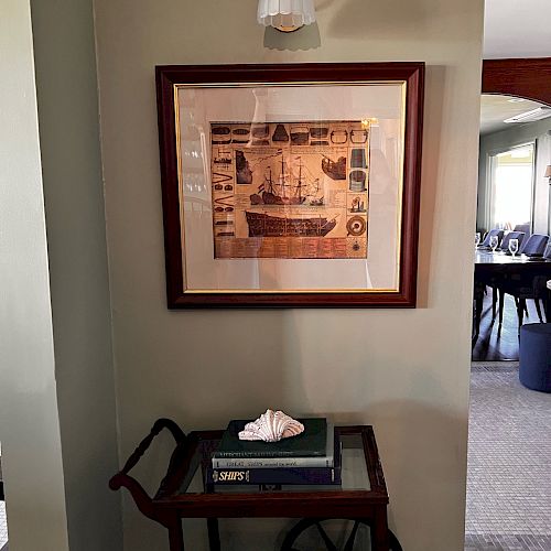 A framed picture of a ship, a small lamp above, and a cart with books and a seashell are placed against a wall in a well-lit room.