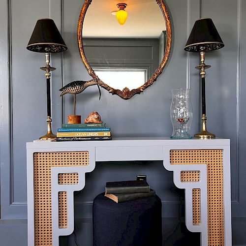 An elegant console table with two black lamps, a decorative bird, and books beneath an ornate oval mirror, all set against a gray wall.