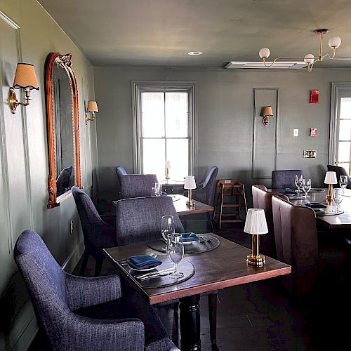 A cozy restaurant interior with tables set for dining, grey upholstered chairs, table lamps, a large mirror on the wall, and two windows allowing natural light.