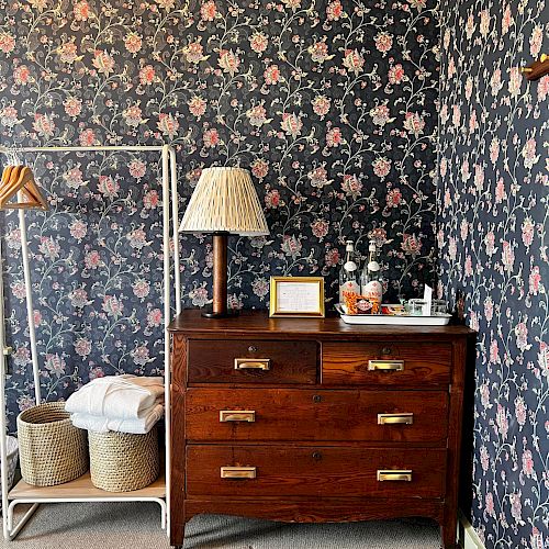 A cozy room with floral wallpaper, a wooden dresser with a lamp and decor, and a standing rack with baskets and clothes.