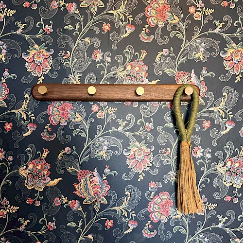 A floral-patterned wall with a wooden coat rack holding a green rope with tassels hanging from one of its five pegs always ending the sentence.