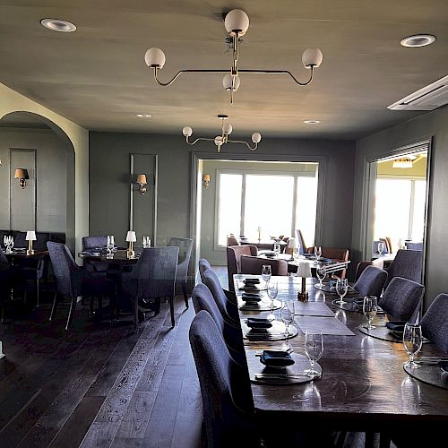 The image shows a modern dining room with dark wood tables, gray chairs, contemporary lighting fixtures, and large windows providing natural light.