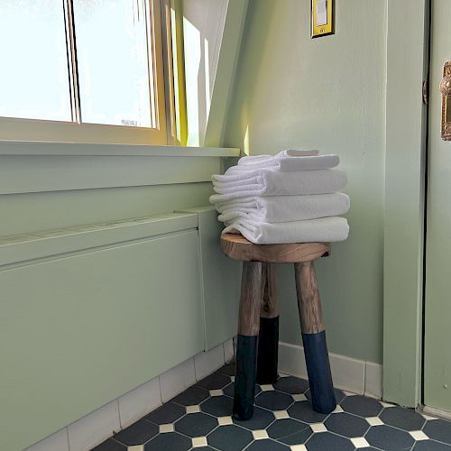 The image shows a stack of folded white towels on a small wooden stool in a bathroom with light green walls and a door.