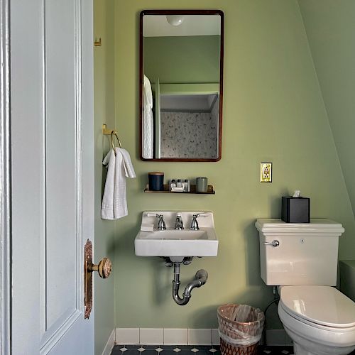 A small bathroom with a green wall, a white sink, a mirror, a towel on a towel ring, a toilet, and a trash bin.