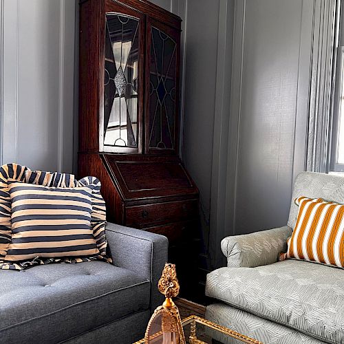 This image features a cozy corner with two armchairs, striped pillows, a wooden cabinet, and a glass coffee table adorned with decorative items.